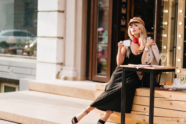 Привлекательная молодая женщина отдыхает после работы в любимом кафе и наслаждается вкусом кофе. Открытый портрет блондинки в стильном наряде, расслабляющемся в выходные.