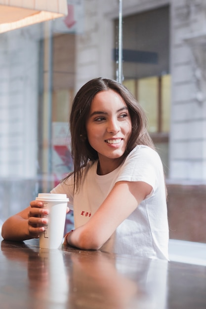 テイクアウトのコーヒーカップを保持しているカフェに座っている魅力的な若い女性