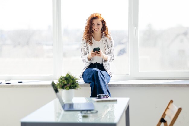 매력적인 젊은 여성이 집 창턱에 앉아 전화로 문자를 주고받는 메시지 전화를 보고 있는 여성은 밝은 미소로 인터넷 현대 스마트폰을 사용합니다.