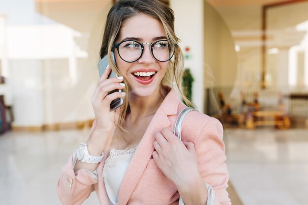 Attractive young woman smiling and talking by phone, looking to the side, standing in hall. She has white short manicure, watches on her wrist. Wearing stylish pink jacket.