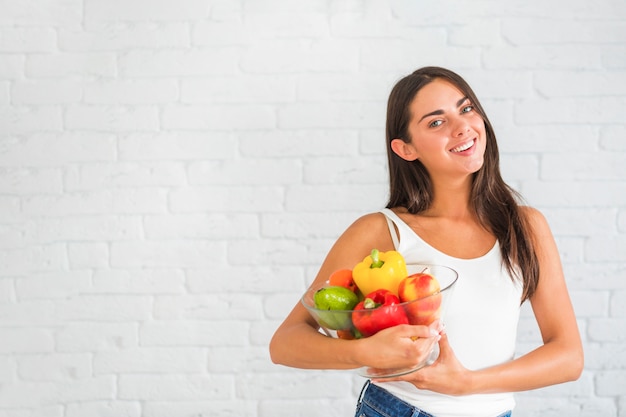 Attractive young woman standing against wall holding bowl of fresh vegetables and fruits