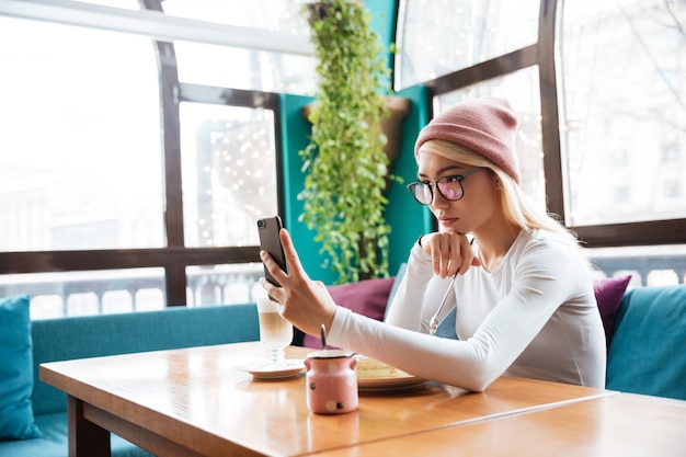 カフェで携帯電話でselfieを取って魅力的な若い女性