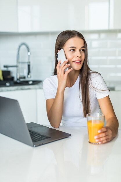 Attractive young woman talking on mobile phone while standing on a kitchen with glass of juice