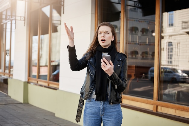 Attractive young woman in trendy leather coat pointing with palm aside while looking up and holding smartphone