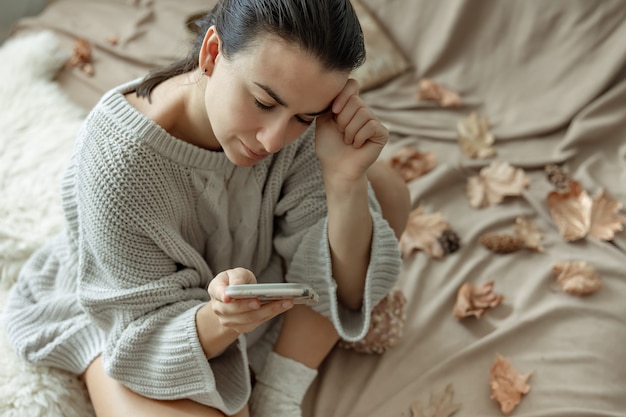 Бесплатное фото Привлекательная молодая женщина пользуется телефоном, сидя в постели среди осенних листьев в уютном вязаном свитере.