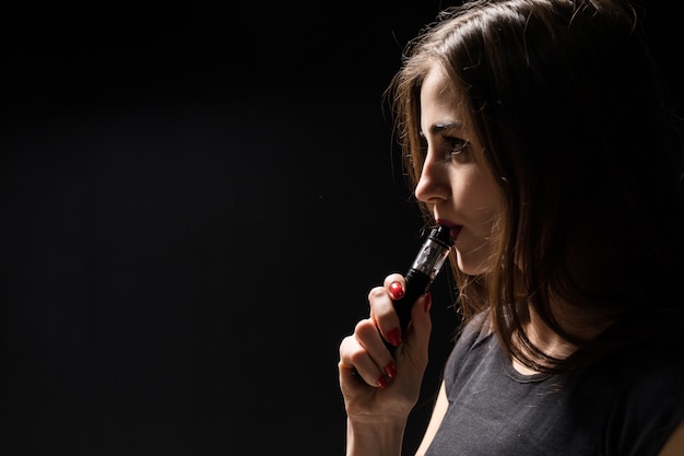 Attractive young woman vaping and blowing smoke isolated on black wall