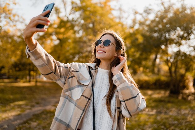 전화를 사용하여 재킷을 입고 가을에 걷는 매력적인 젊은 여자