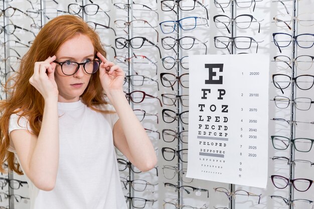 Очки привлекательной молодой женщины нося стоящую аккуратную диаграмму snellen в optica