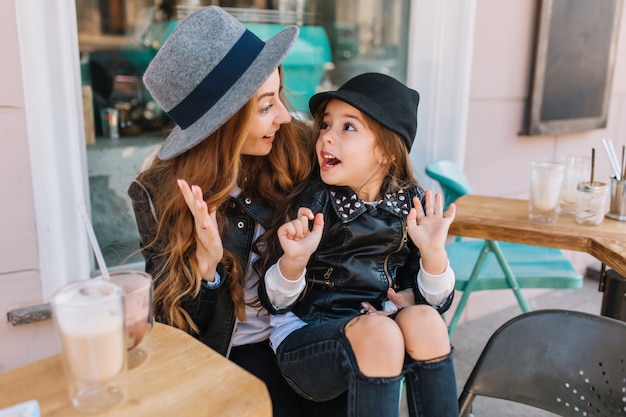 Attractive young woman wearing gray hat fooling around with cute daughter after drinking milk shake.