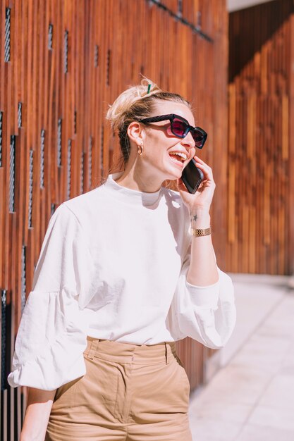 Солнечные очки привлекательной молодой женщины нося смеясь над пока принимающ на мобильный телефон