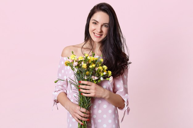 黒の長いウェーブのかかった髪を持つ魅力的な若い女性は、水玉のドレスに身を包んだ花を保持し、春の気分、淡いピンクのポーズ、ボーイフレンドとのロマンチックなデートをしています。 3月8日のコンセプト