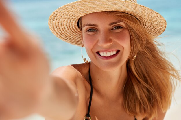 넓은 미소, 건강한 피부를 가진 매력적인 젊은 여성이 해변에 달려 있고, 좋은 분위기에있는 자신의 사진을 찍고, 여가와 여름 휴가를 즐깁니다. 아름다운 여성은 바다에 셀카를 만든다
