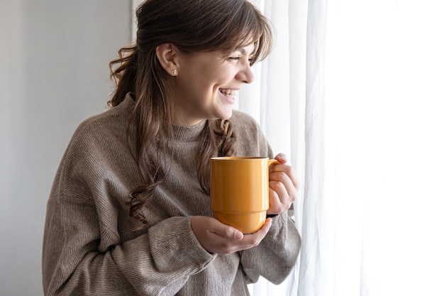 窓の近くに温かい飲み物のカップを持つ魅力的な若い女性