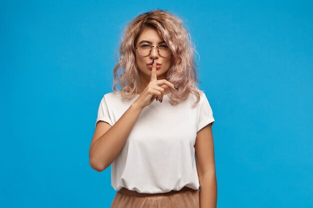 Attractive young woman with curly messy hair wearing round eyeglasses and oversize top asking to keep silent, holding fore finger at her lips, making shushing sound. Secrecy