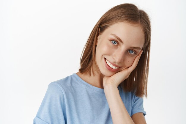 Attractive young woman with fair short hair and smooth, glowing natural skin, touch her face and smiling white teeth, standing against white wall