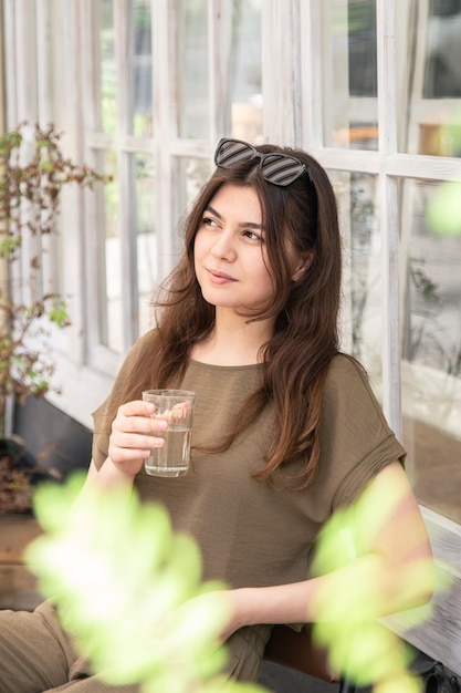 カフェのテラスで夏の日にコップ一杯の水を持つ魅力的な若い女性