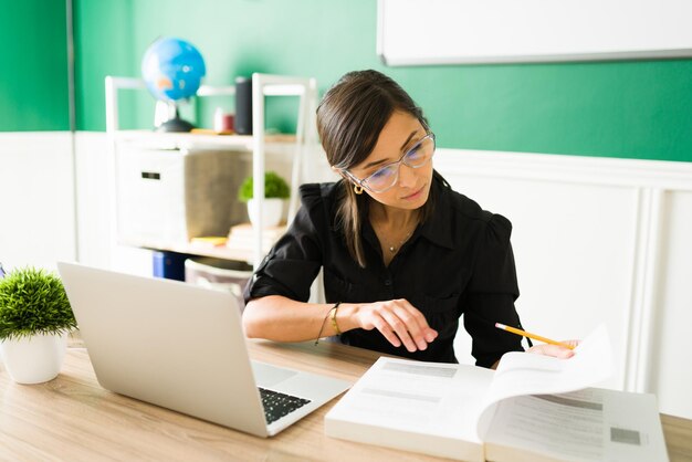 自宅で教科書と彼女のラップトップで彼女のクラスを計画しながら勉強している眼鏡をかけた魅力的な若い女性