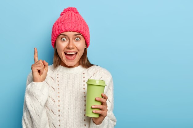 Attractive young woman with joyful face expression, points fore finger above, demonstrates cool promotion, holds green takeaway cup of coffee, wears winter outfit