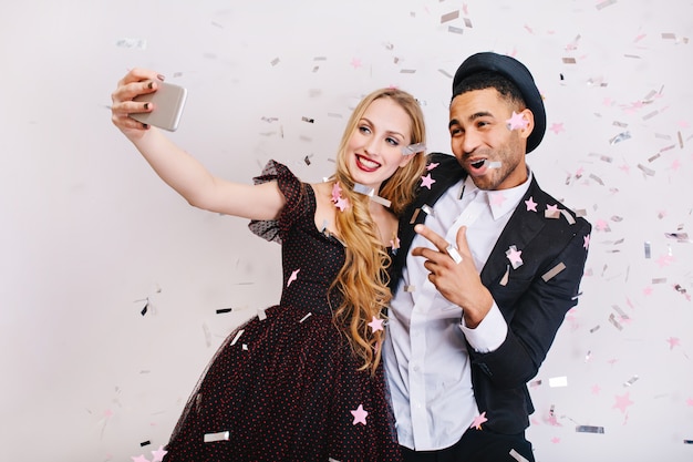 Attractive young woman with long blonde hair in luxury evening dress making selfie in tinsels with joyful handsome man. Celebrating party