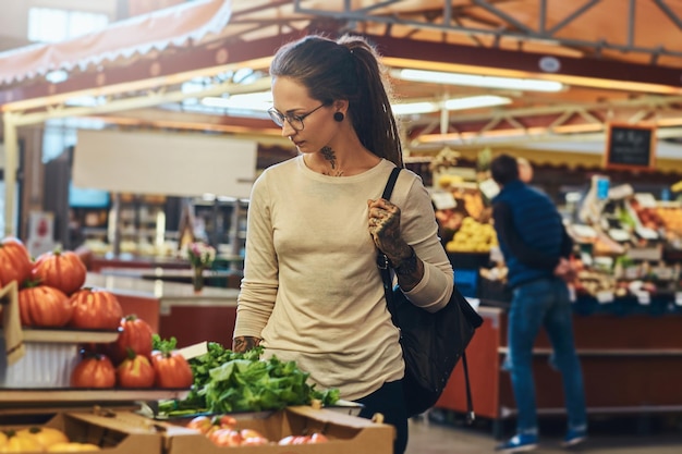 Foto gratuita giovane donna attraente con il tatuaggio e gli occhiali che visitano il mercato locale per le verdure fresche.