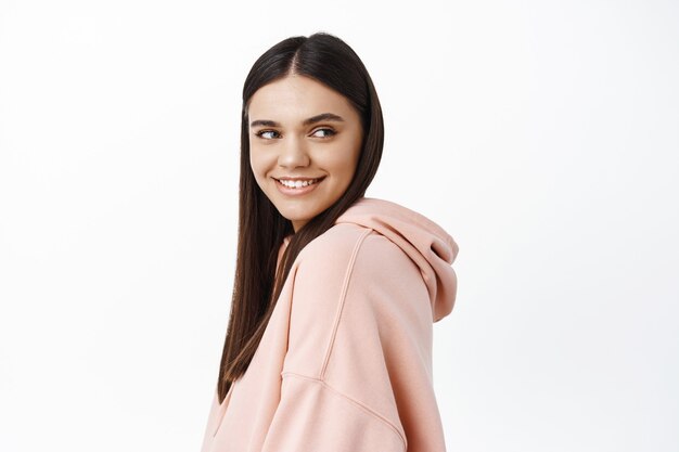 Attractive young woman with white smile teeth, turn head at right side copy space with pleased and confident face, standing in hoodie against white wall