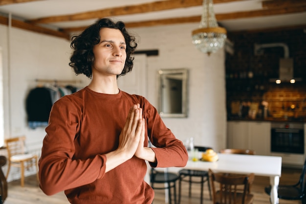 Attracttive joyful young man holding hands pressed together on his chest expressing gratitude, making namaste gesture