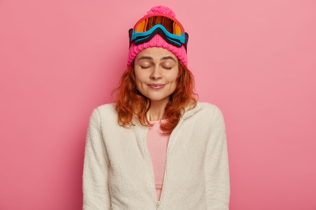 Attractve redhead woman wears warm hat, ski goggles and white jumper