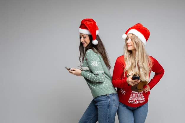 Притягательные девушки-друзья в красно-белых новогодних шапках с телефонами