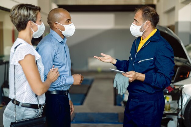 Auto mechanic and a couple wearing face masks while talking at car service workshop