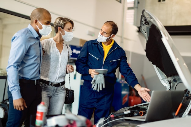 Auto mechanic and his customers talking while examining vehicle breakdown in a workshop during coronavirus pandemic