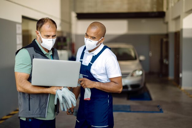 Auto mechanics with protective face masks using laptop in repair shop