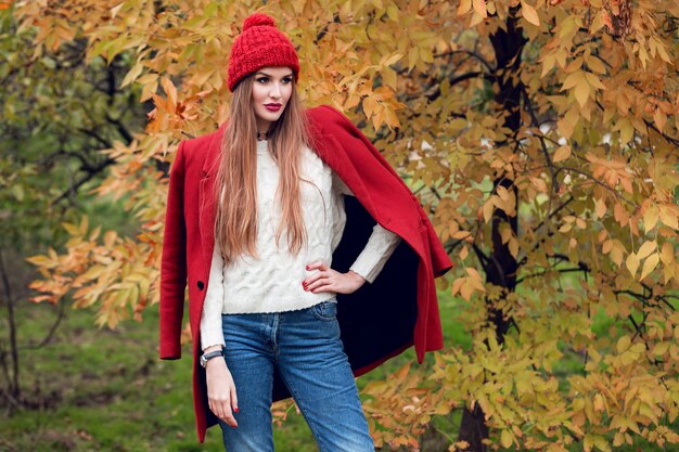 Autumn fashion portrait of blonde woman in red stylish coat and knitted hat walking in park.
