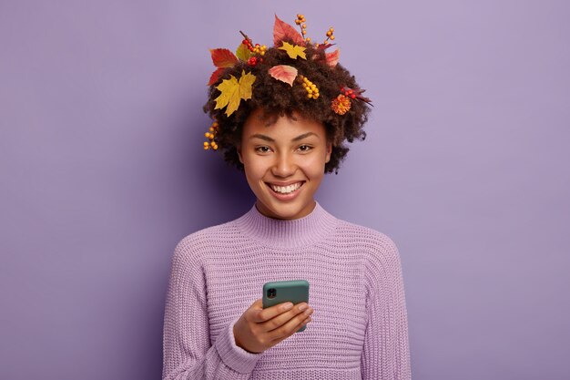 Осень, концепция технологий. Счастливая афроамериканка пользуется современным смартфоном, радостно улыбается, у нее вьющиеся волосы, украшенные листвой