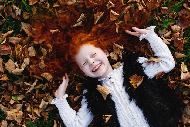 Осенние флюиды, детский портрет. Очаровательная и рыжеволосая маленькая девочка выглядит счастливой, лежа на опавшем леве
