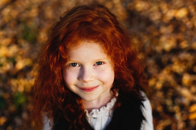 Autumn vibes, child portrait. Charming and red hair little girl looks happy standing on the fallen l