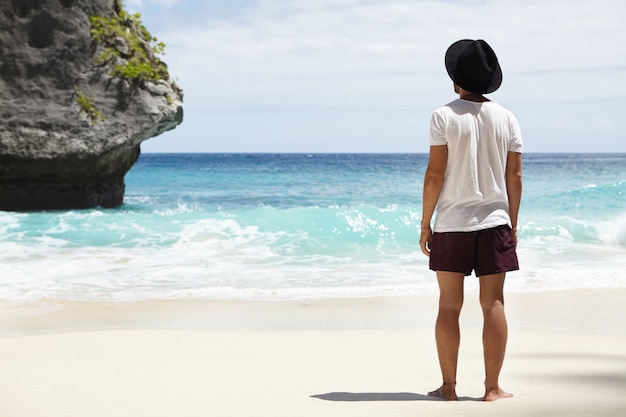 観光コースから離れて。ターコイズブルーの海の石の島の前の砂浜に立っている裸足の若い白人の冒険家