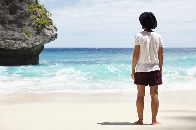 Away from tourist trails. Barefooted young Caucasian adventurer standing on sandy shore in front of stone island in turquoise ocean which he finally found during his long journey along coastline