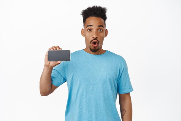 Awesome app check it out. Amazed african american guy gasp, say wow, showing horizontal smartphone screen, mobile phone application interface, staring excited at camera, white background.