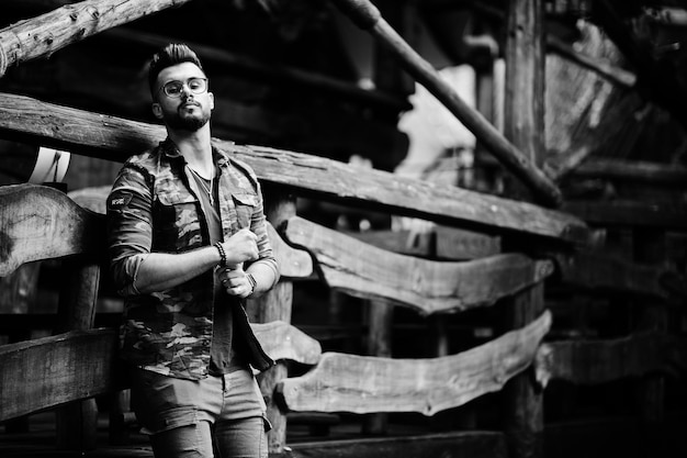 Awesome beautiful tall ararbian beard macho man in glasses and military jacket posed outdoor against wooden rustic house