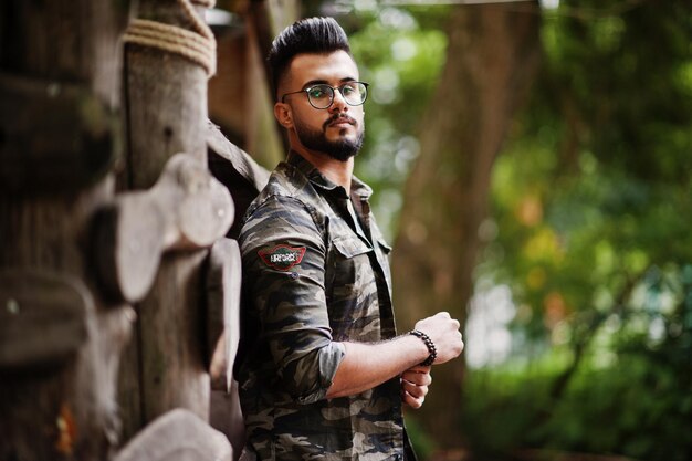 Awesome beautiful tall ararbian beard macho man in glasses and military jacket posed outdoor against wooden rustic house