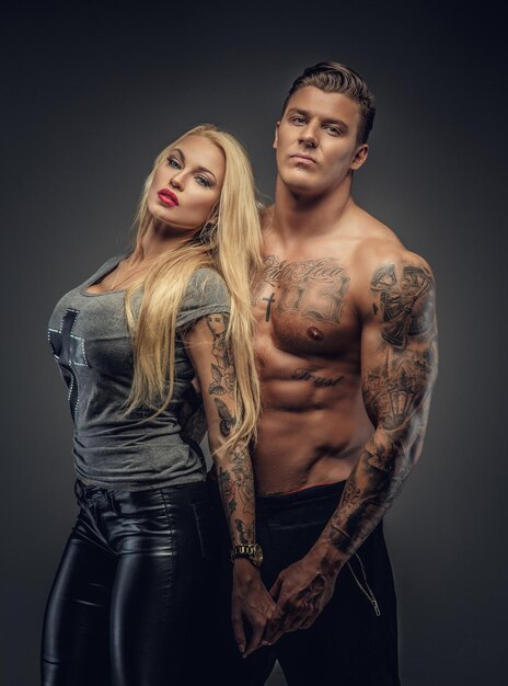 Awesome blond woman posing with shirtless muscular tattooed man. Isolated on grey background.