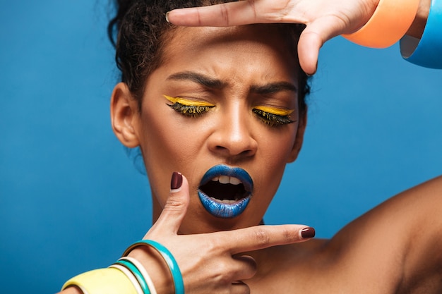 Awesome portrait of emotional afro american woman with bright makeup and accessories covering face with hands, over blue wall