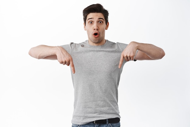 Awesome promo here. Impressed adult guy look surprised, pointing fingers down at cool deal, showing banner, standing over white wall