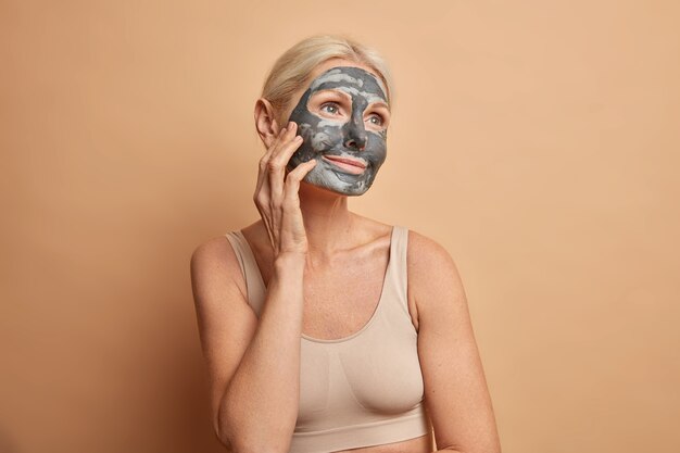Awesome relaxed woman applies clay mask on face touches cheek and looks with dreamy expression has natural beauty undergoes cosmetic procedures dressed in cropped top isolated on beige wall