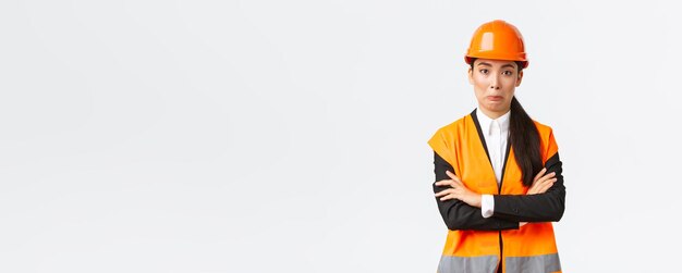 Awkward asian female engineer in reflective jacket safety helmet cross arms chest and smirk surprised making silly mistake acting natural standing white background Copy space