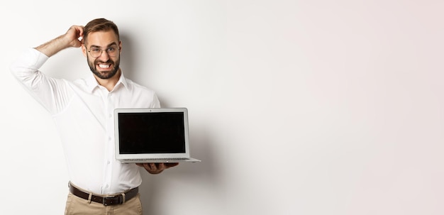 Foto gratuita uomo d'affari imbarazzante che mostra lo schermo del laptop e che sembra dubbioso in piedi su sfondo bianco onu