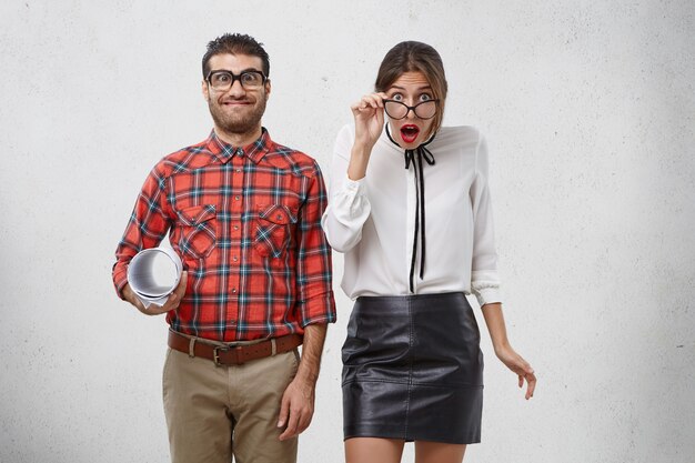 Awkward cheerful man with comic expression, holds blueprint, stands near shocked surprised female