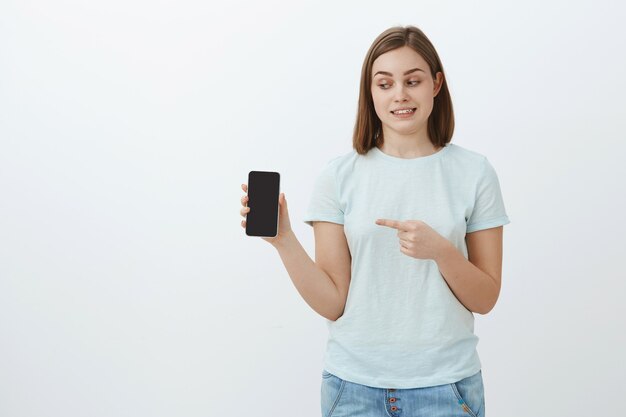 awkward cute female student in trendy t-shirt and jeans holding smartphone pointing at phone screen while showing strange photo of friend standing over gray wall
