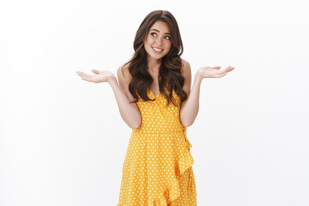 Awkward reluctant young brunette in yellow dress, look away embarrassed shy, shrugging and spread hands sideways