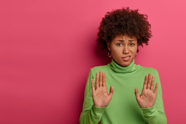 Awkward unimpressed woman with Afro hairstyle, pulls palms towards camera, refuses something, rejects proposal, wears green neck sweater